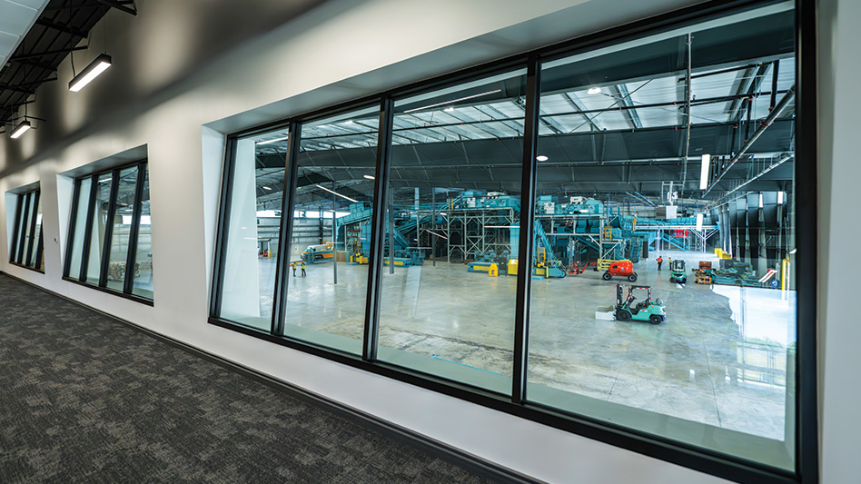 A view of the inside of a recycling facility window overlooking workers moving around the floor of the MRF.