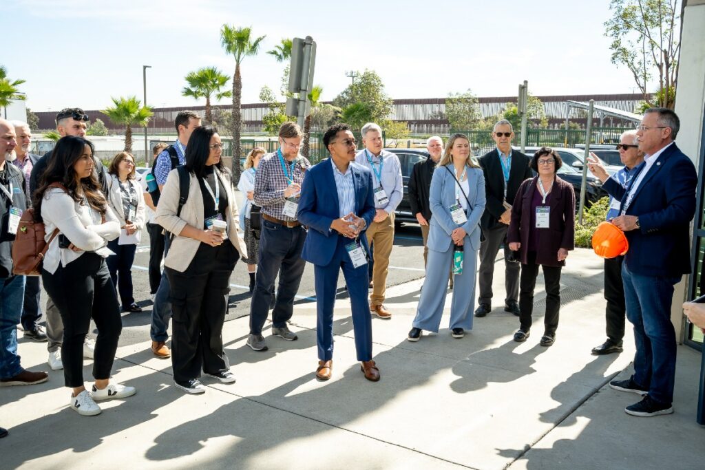 Group of professionals outdoors listening to a man holding an orange hard hat during a site tour.