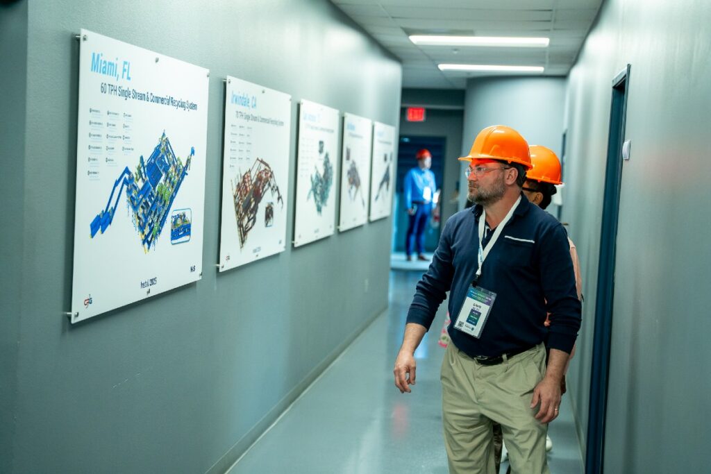 Man in orange hard hat and badge walking down a corridor, looking at technical posters of machinery mounted on the wall.