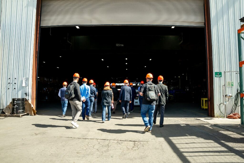 Group of people wearing orange hard hats walking into a large open warehouse entrance.