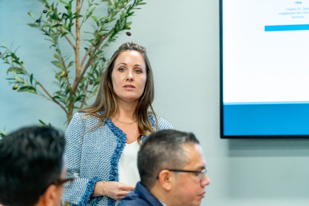 Woman in light-blue tweed jacket standing and presenting in a meeting room beside a screen, colleagues blurred foreground.