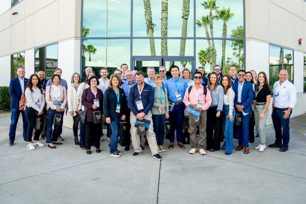 Large mixed-age group of conference attendees in business casual posing outside a glass building with palm trees reflected.