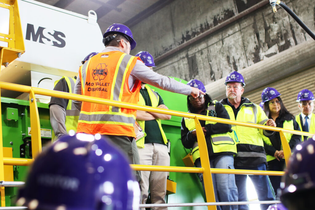 Foreman in orange vest points as workers in purple hard hats and yellow safety vests stand on a yellow railing in a facility.