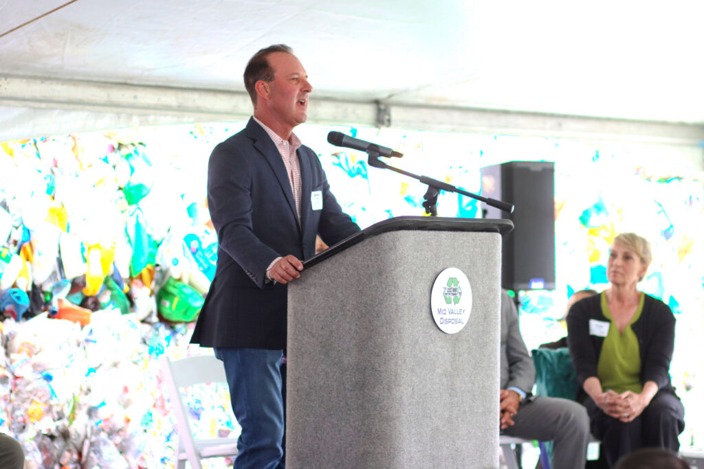 Man in navy blazer speaking at a podium with a microphone in front of a colorful recycling-materials backdrop.