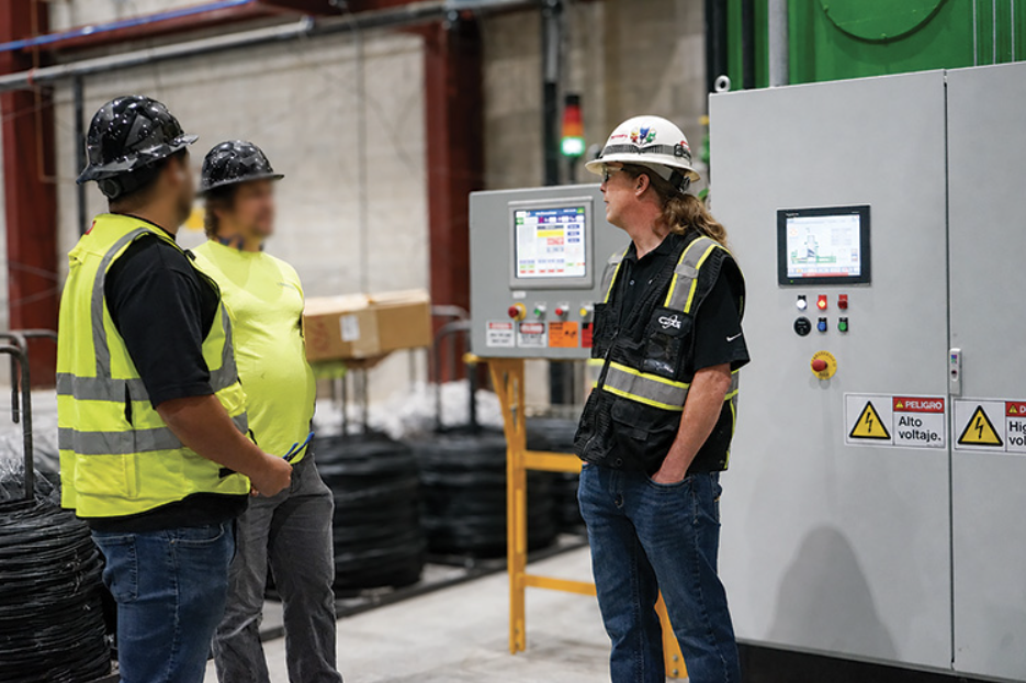 Three workers in hard hats and high-visibility vests talk beside industrial control panels with voltage warning signs.