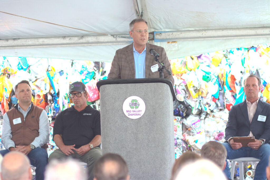 Speaker in a plaid jacket at a podium labeled Mid Valley Disposal with colorful recycled-plastic bales behind him.