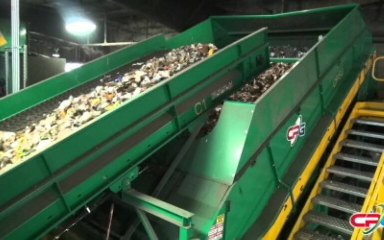 This image shows a large green industrial conveyor belt system transporting mixed waste materials for sorting or processing in a recycling or waste management facility.