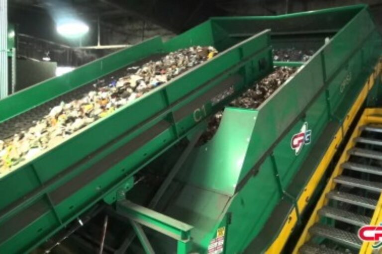 This image shows a large green industrial conveyor belt system transporting mixed waste materials for sorting or processing in a recycling or waste management facility.