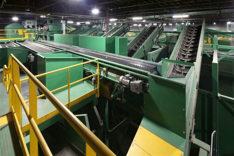 A large indoor industrial facility with green and yellow machinery and conveyer belts. Multiple conveyer systems with metal railings are seen, alongside various interconnected equipment for material processing or sorting. The background is dimly lit.