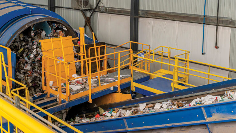 A recycling facility with a large Trommel machine sorting and processing mixed recyclable materials. Yellow safety railings surround the metal structure, and the conveyor belt carries items to be sorted. The background shows a high ceiling and industrial interior.