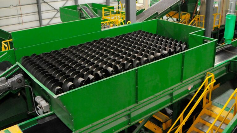 A large CP Auger Screen machine with multiple black rotating cylinders is shown in a factory setting. The machine appears to be part of a recycling or sorting facility, with yellow railings and stairways around the equipment for access and maintenance.