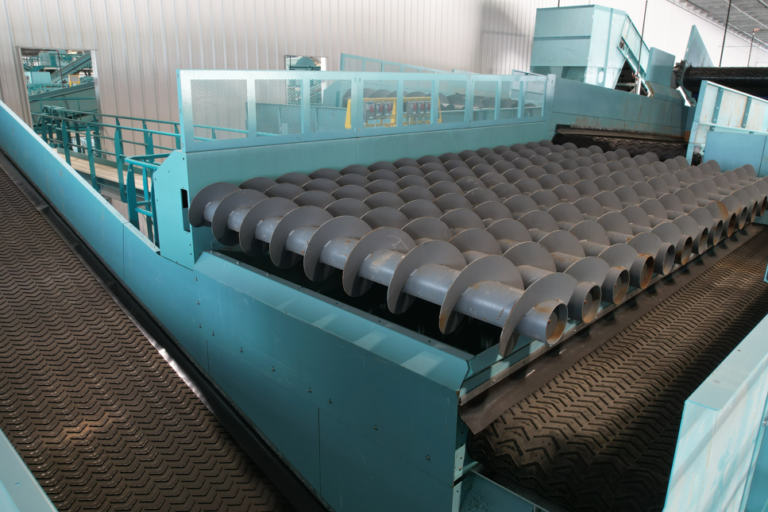 A large CP Auger Screen system is shown, featuring a series of rotating cylindrical rollers. The machinery is painted in turquoise and is situated in a factory setting with metal guardrails and an industrial ceiling overhead.