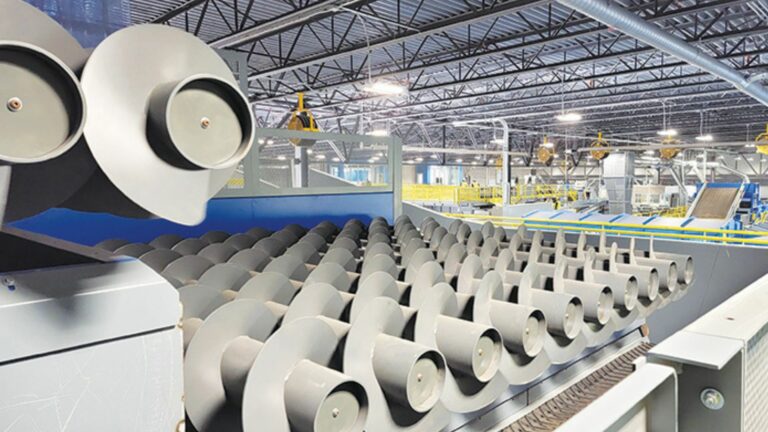 A modern MRF facility featuring a large, gray metal CP Auger Screen for processing materials. The background reveals numerous conveyor belts, machinery, and structural components under a ceiling supported by metal beams.