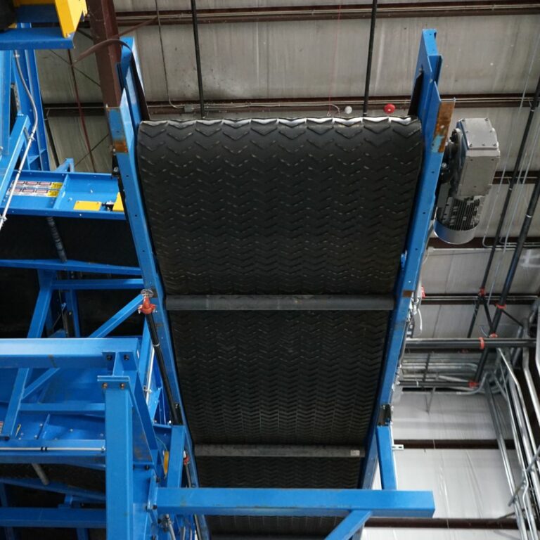 A large, industrial conveyor belt system viewed from below, displaying a black, rubberized belt with a textured pattern. The machinery is framed by blue metal supports and situated within a warehouse environment, with electrical wiring visible on the ceiling.