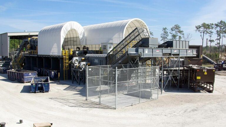 Industrial recycling facility with large white, arch-shaped structures and various equipment like conveyor belts. Metal fences surround some parts, and several large containers are scattered around. Trees and a clear blue sky are visible in the background.