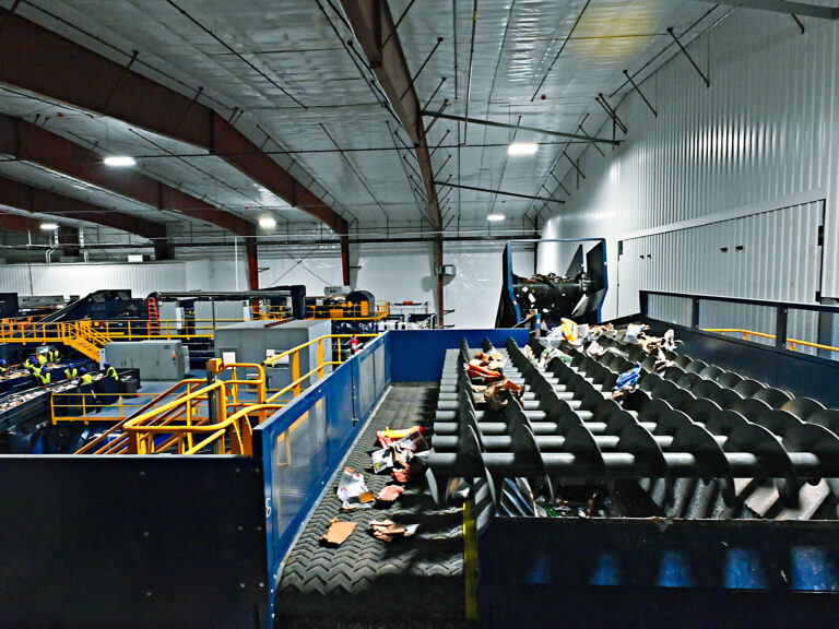 A large indoor recycling facility with sorting machinery. The CP Auger Screen is lined with various recyclable materials such as paper and plastic. Industrial lighting illuminates the spacious area, with metal beams supporting the ceiling. Workers in safety vests are seen in the background.