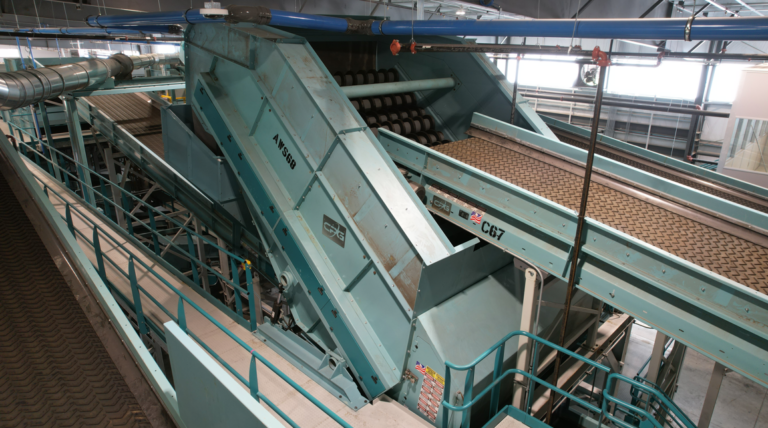 A large CP Screen machine with multiple conveyor belts and metal structures is seen in a factory or processing plant. The machinery appears to be used for material handling or sorting, with various paths and components leading in different directions.