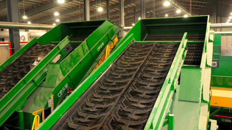 A close-up of a CP Disc Screen in a MRF setting. The large green belts are angled upwards, with rubberized gripping surfaces. The background shows a well-lit, high-ceilinged industrial space with metal beams and other machinery.