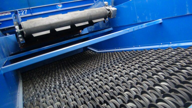 Close-up image of a CP Disc Screen machinery featuring a conveyor belt with a black, rubberized surface. The machine has blue metallic components and appears to be part of a sorting or processing system.