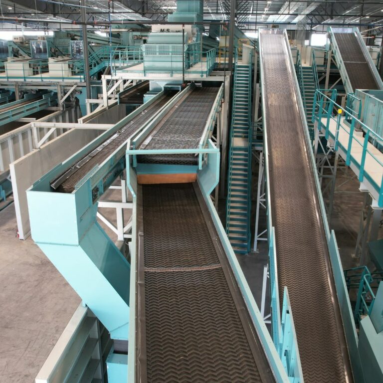 An industrial facility with several interconnected conveyor belts and elevated walkways. The metal belts and teal-colored machinery create a complex network for transporting materials. Overhead lighting brightens the expansive space.