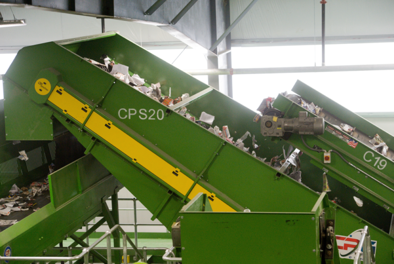 CP Disc Screen labeled "C19" and "CPS20" in a recycling facility transport mixed recyclable materials. The belts are green with yellow accents and carry various types of paper, plastic, and other items. The background shows industrial equipment and a lit interior.