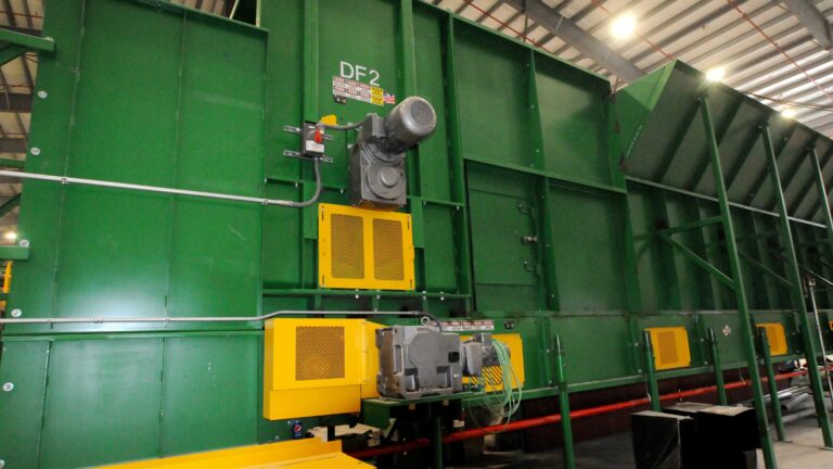 A large green Drum feeder machine with yellow components. The machine has various cables and a gray motor on its side. The setting appears to be inside a factory or warehouse with a high ceiling and metal beams.