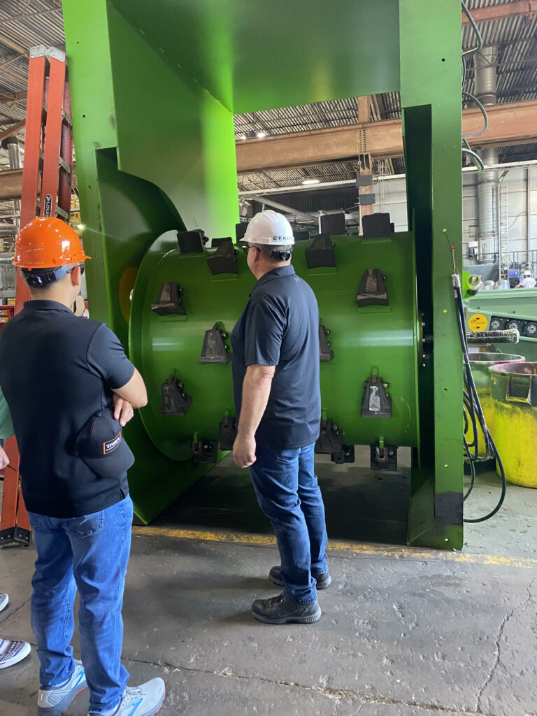 Two men in safety helmets stand in front of a large green Drum feeder machine. One wears an orange helmet, and the other wears a white one. They appear to be inspecting or discussing the equipment inside a factory with various tools and machinery in the background.