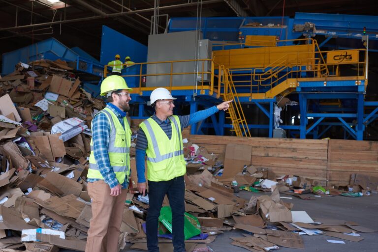 Two people wearing safety vests and helmets stand in a recycling facility surrounded by piles of assorted recyclable materials. One person points towards a large machine in the background as they discuss the operations. Workers and equipment are visible in the background.