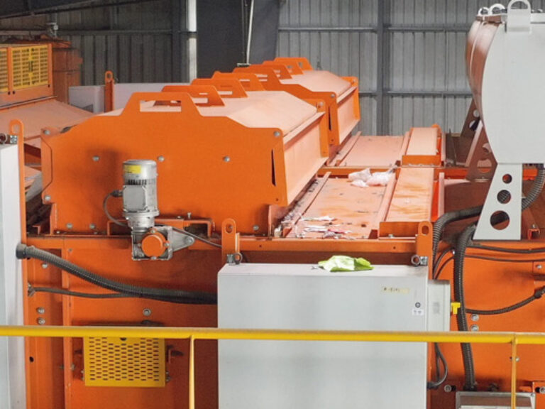 A panoramic view of an Optical Sorter machinery setup with orange-painted equipment, control panels, and a "MSS" label on one section. The machinery is enclosed by a yellow safety barrier, with various pipes and cables visible in the background.