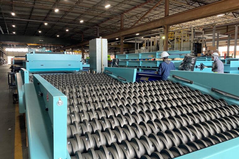 A large industrial facility with a glass breaker system consisting of numerous rollers. Several workers wearing helmets and protective clothing are inspecting and working on the machinery. The spacious facility is well-lit with overhead lighting.