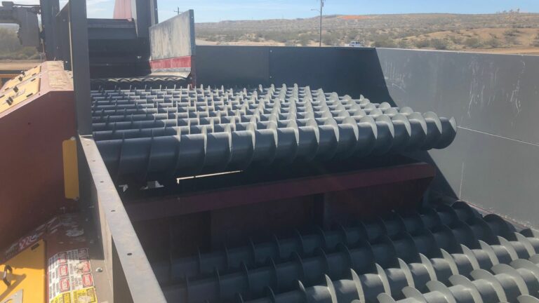 Conveyor belts with cylindrical Auger rollers in an industrial setting under a blue sky. The equipment is designed for transporting or processing materials. In the background, there are some hills and sparse vegetation.