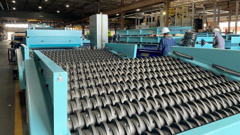 A large industrial facility with a glass breaker system consisting of numerous rollers. Several workers wearing helmets and protective clothing are inspecting and working on the machinery. The spacious facility is well-lit with overhead lighting.