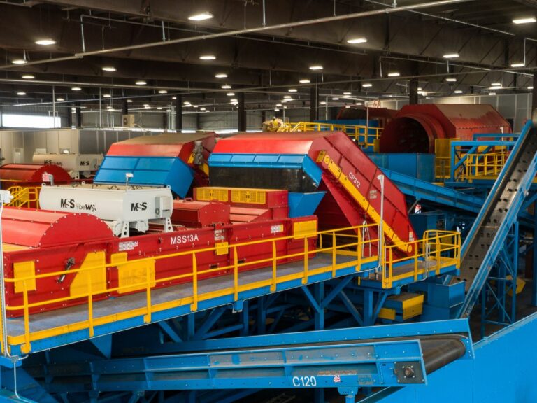 A large, brightly lit industrial MRF facility houses several red, yellow, and blue machines used for materials sorting or processing. Conveyor belts and walkways weave through the machinery, and a few workers are visible in the background wearing safety gear.