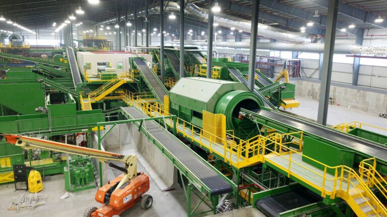 A large indoor MSW recycling facility with various green machinery and conveyor belts. A rotating drum and multiple sorting stations are visible. An orange boom lift is parked in the foreground. The setting is well-lit and spacious, with a high ceiling.