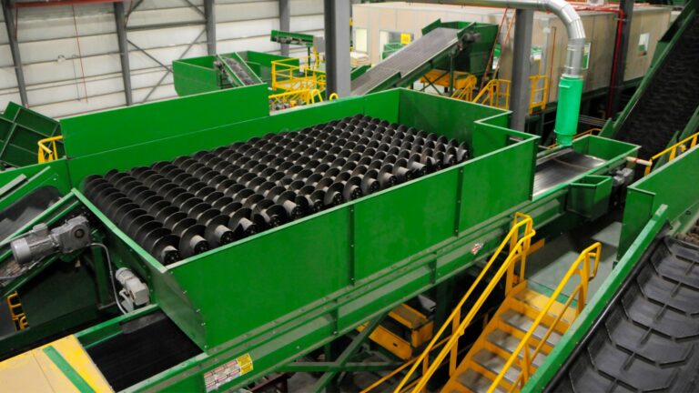 A large industrial Auger sorting machine in a recycling plant. The machinery is predominantly green and features conveyor belts and rows of black cylindrical rollers. Yellow safety railings line the pathways around the equipment.