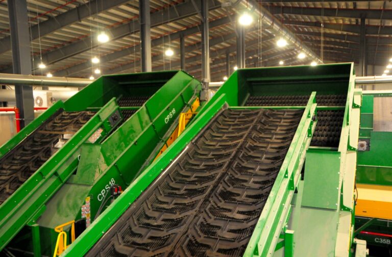 An overhead view of an industrial recycling facility featuring green machinery with conveyor belts used for sorting and processing materials. The equipment is surrounded by a network of platforms, chutes, and walkways.