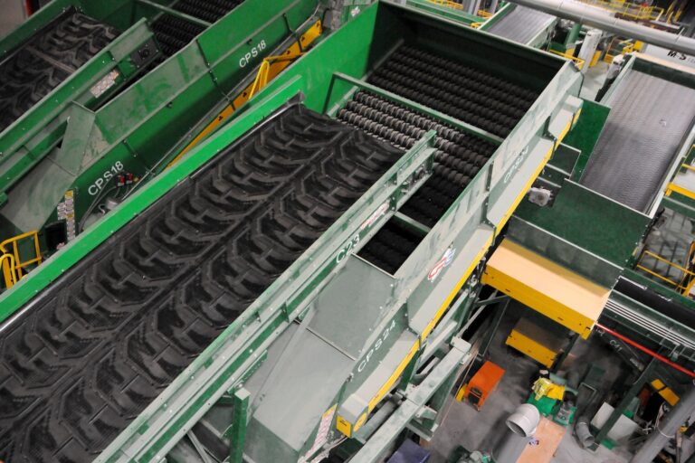 An overhead view of an industrial recycling facility featuring green machinery with conveyor belts used for sorting and processing materials. The equipment is surrounded by a network of platforms, chutes, and walkways.