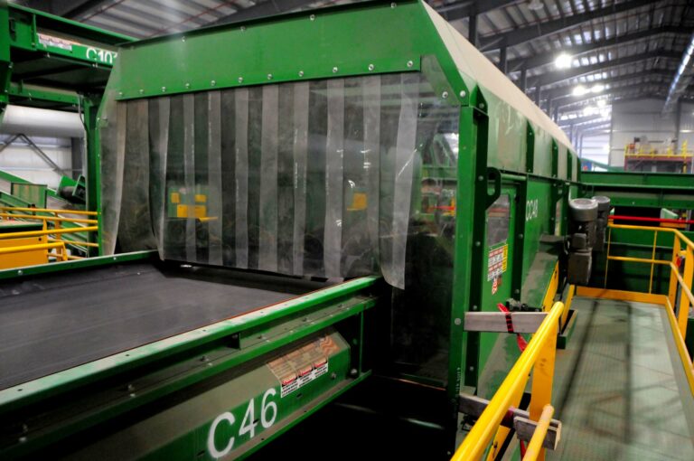 A green OCC Screen machine with conveyor belts inside a well-lit warehouse. The machine is labeled "C46" and features a protective transparent barrier with vertical slats. Yellow safety railings surround the machinery.