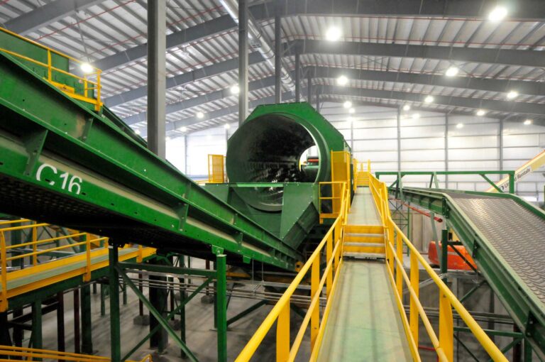 A large indoor waste management facility with various green machinery and conveyor belts processing materials. A cherry picker is positioned on the ground below, and bright overhead lights illuminate the space. The facility has high ceilings and an industrial design.