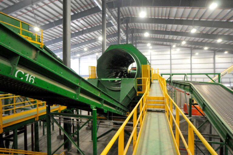 A large industrial facility with green machinery, including conveyor belts and a cylindrical rotating drum. The equipment is elevated with yellow safety rails, and the interior of the building has a high ceiling with supportive steel beams.