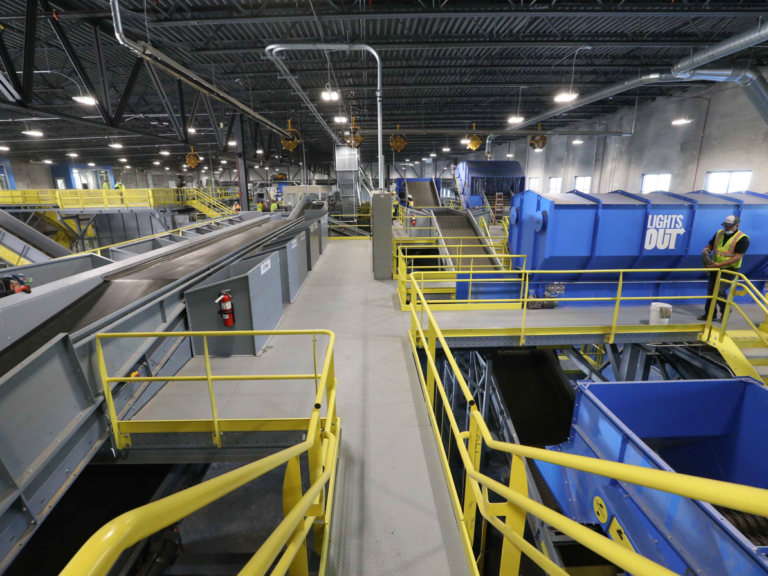 A spacious recycling facility with multiple conveyor belts, guardrails, and machinery. Workers in high-visibility vests are operating the equipment under large overhead lights. The facility has a blue container labeled "LightsOut"