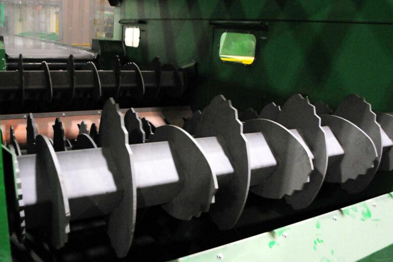 A close-up view of OCC Screen machinery featuring multiple large, grey, screw-like augers inside a green housing. The augers have serrated edges and are designed to process or move material within the equipment.