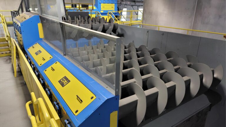 A large OCC Auger machine with multiple spiral-shaped metal components. The machine is predominantly blue and yellow, and is situated in a MRF setting with gray walls and yellow safety railings.