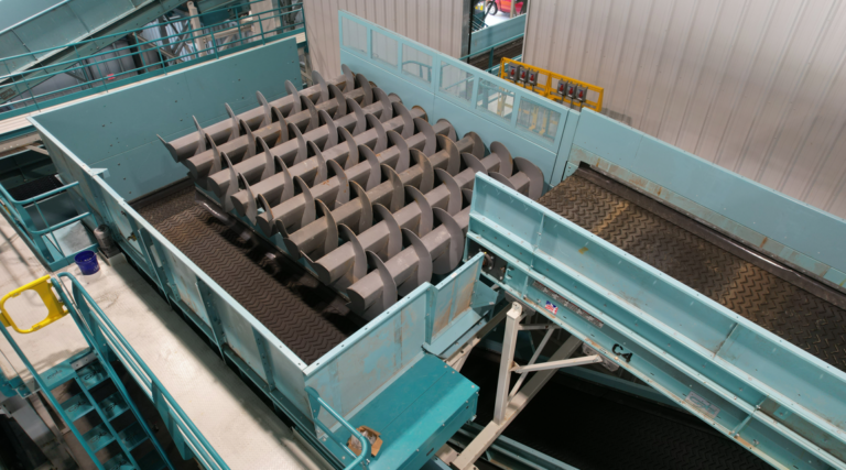 An OCC Auger Screen system painted in teal, comprised of multiple tracks and large rotating metal screw-like mechanisms, used for processing or transporting materials in a facility. The system is situated indoors, surrounded by metal railings and walkways.