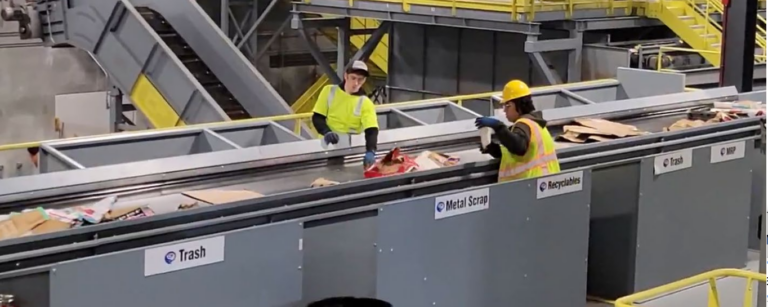 Two workers wearing high-visibility safety vests and helmets are sorting recyclable materials on a conveyor belt in an industrial facility. Various pieces of cardboard and other recyclables are seen on the belt. Industrial machinery and staircases are in the background.