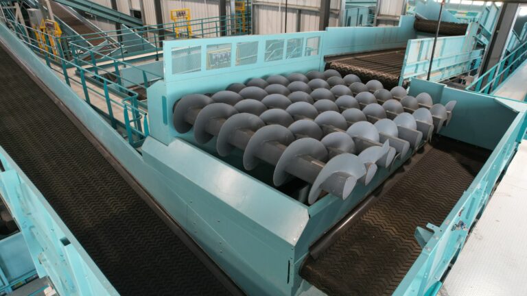 Industrial machinery consisting of multiple large, curved metal auger rollers aligned in a row, likely used for processing waste materials. The equipment is housed in a facility with metal walkways and railings around it.