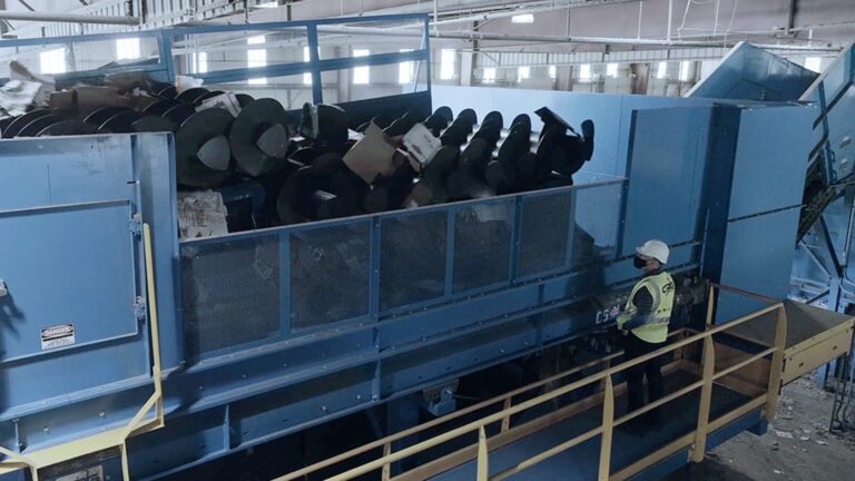 A worker wearing a hard hat and safety vest stands next to a large blue Auger machine filled with waste. The machine is situated in a spacious warehouse with windows and overhead lights.