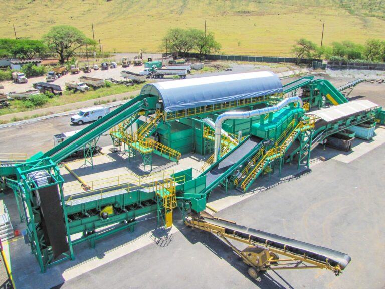 An expansive industrial recycling facility features green conveyor belts, metal structures, and a large covered area. Several workers and vehicles are visible in the surrounding area. Trees and a grassy field can be seen in the background.