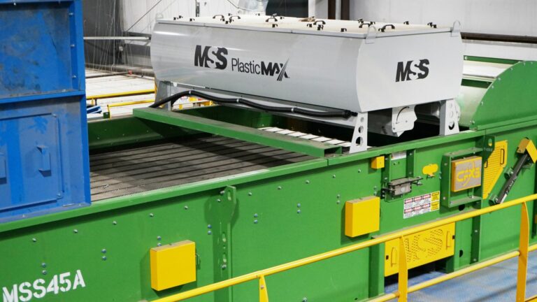 An automated material sorting machine in a recycling plant. The machine, labeled "MSS" and "PlasticMAX," features a conveyor belt and sorting equipment. The surrounding area is industrial with metal platforms and safety railings.