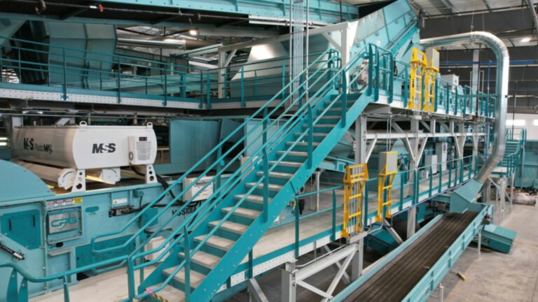 A large MRF facility featuring teal-colored machinery and conveyor belts. Central to the image is a metal staircase leading to an elevated platform. The area is filled with various equipment, pipes, and structures, presumably for material processing or sorting.